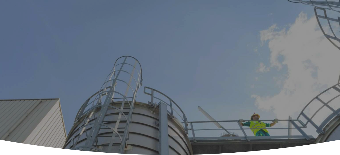 Un lavoratore in abbigliamento da lavoro, con casco di sicurezza giallo e giubbotto ad alta visibilità verde, è in piedi su una piattaforma metallica circolare in cima a un serbatoio industriale. La piattaforma è dotata di corrimano e scale a chiocciola. Lo sfondo mostra un cielo azzurro parzialmente coperto da nuvole bianche.